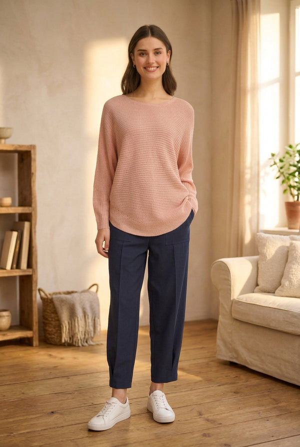 A woman modeling a light pink ribbed knit sweater and navy blue cropped trousers, standing against a plain light gray studio background.