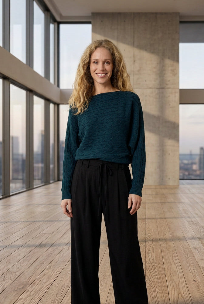 A woman wearing a dark teal blue cable-knit off-the-shoulder sweater and black trousers, standing against a plain light gray wall.