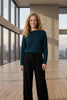 A woman wearing a dark teal blue cable-knit off-the-shoulder sweater and black trousers, standing against a plain light gray wall.
